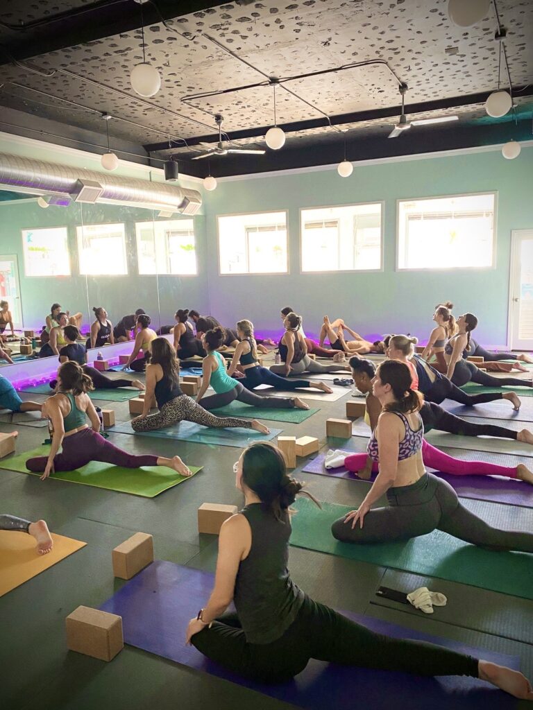 A large indoor yoga class with participants in pigeon pose, using blocks and props on colorful mats in a mirrored studio with ambient lighting.