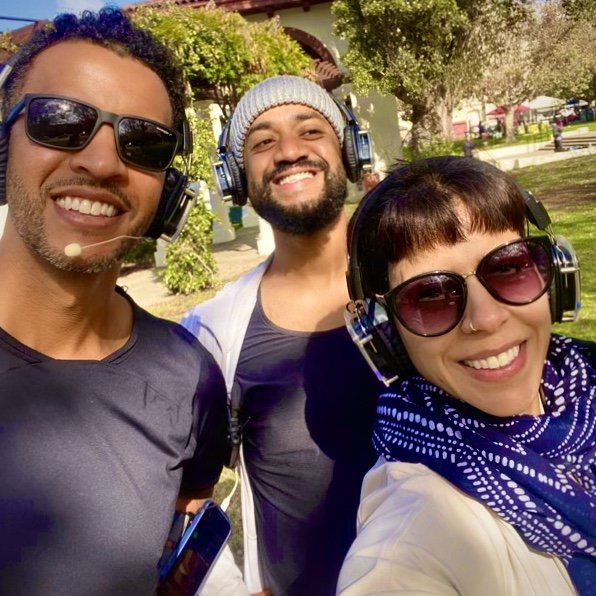 Three smiling people outdoors wearing wireless headphones and sunglasses, participating in what appears to be a silent yoga or meditation event.